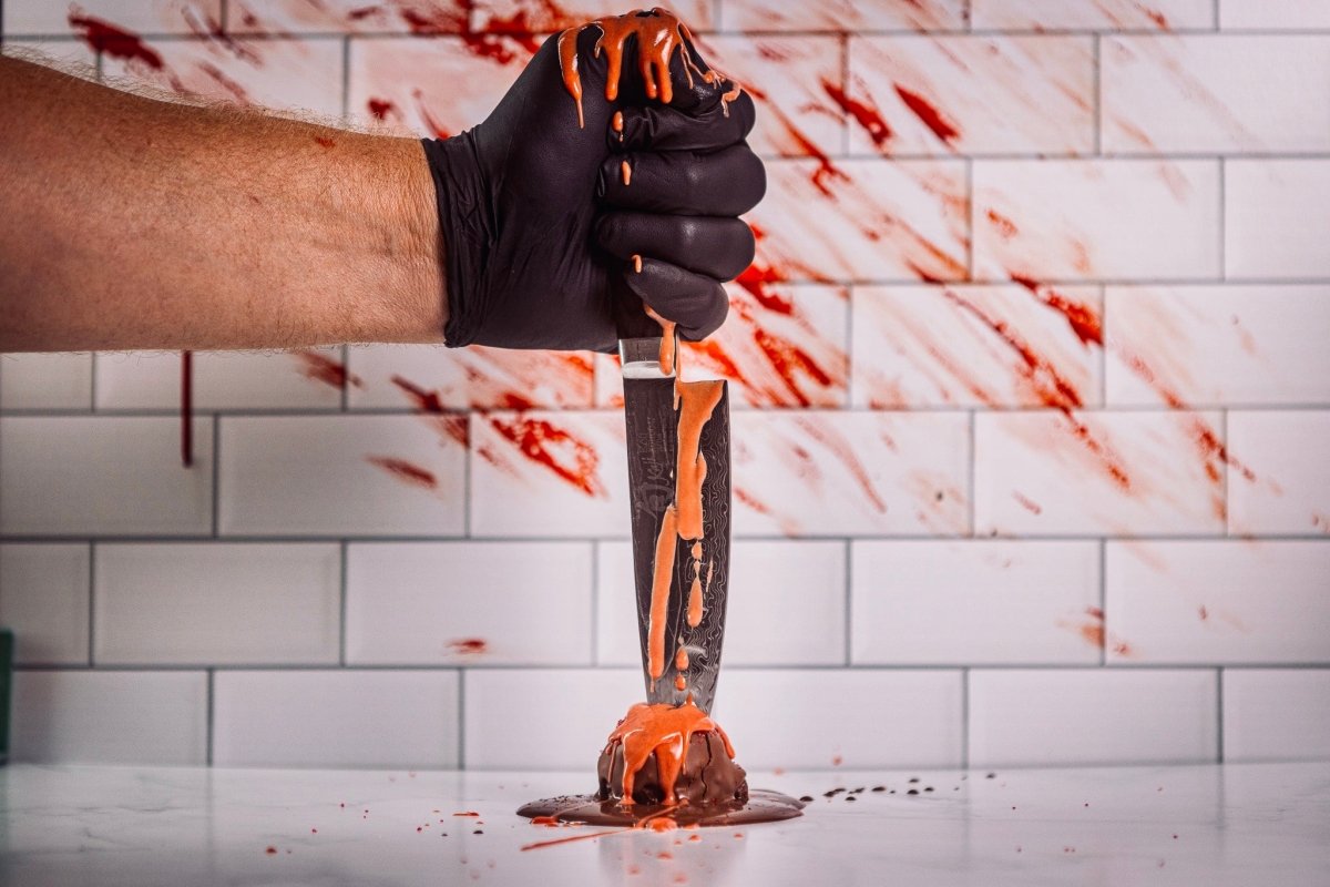 Hand wearing a black glove holding a knife stabbing Chocolate protein truffle coated in rich dark chocolate, against a tiled wall background.